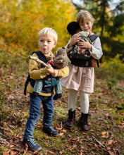 2 children playing outdoors wearing doll carriers with Teddys and dolls in them