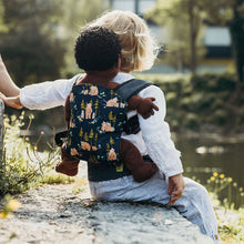 child sitting on a path looking towards a creek wearing a doll carrier on their back with a doll 