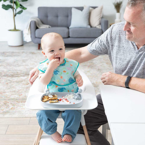 Baby sitting in a white highchair wearing a bib eating food off the tray with and adult sitting beside them