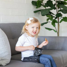 a young girl dress in a white top and blue jeans blonde hair and a white bow clip sitting on a grey couch eating snacks out of a reusable small snack bag 