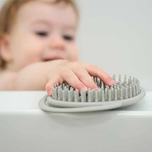 a young child blurred in the background in a bath reaching for a grey silicone brush