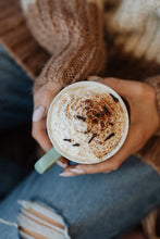 a close up of a woman wearing jeans and a knitted jumper holding a green cup of hot chocolate with whipped cream and chocolate sprinkled on top