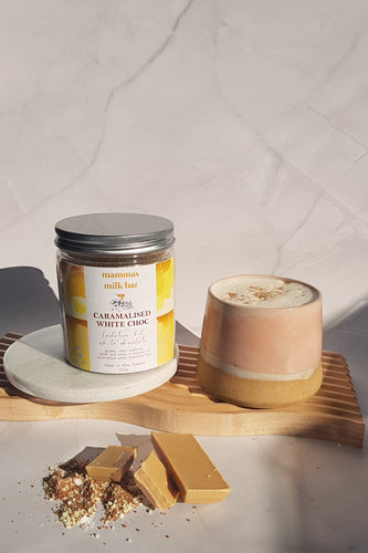 a peach pottery cup filled with a white hot chocolate with the container beside it both on a wooden board, a blend powder and piece of caramel chocolate are sitting in front of it