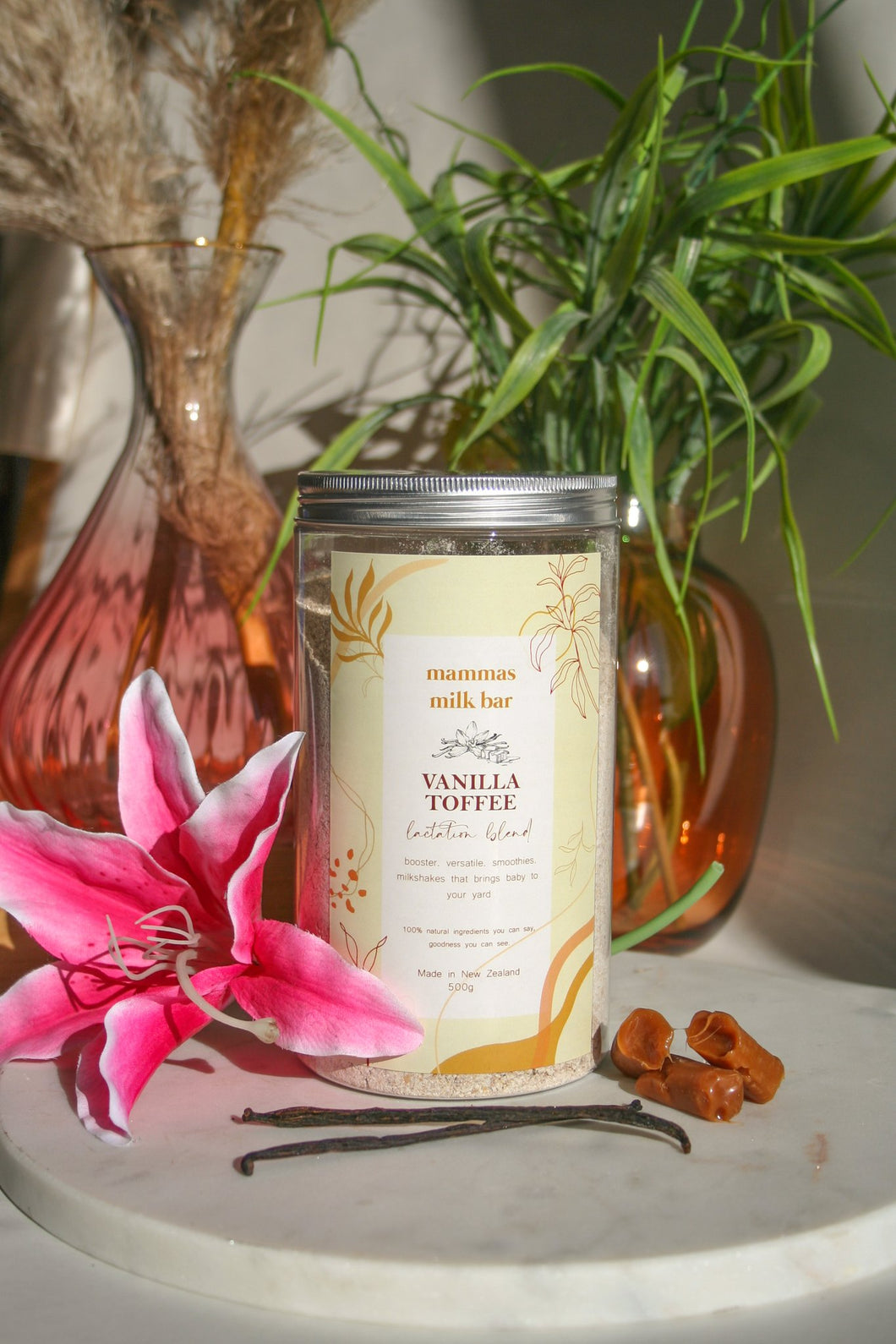 pink lilly, vanilla beans, toffee in from of a large jar of powder, plants in vases behind 