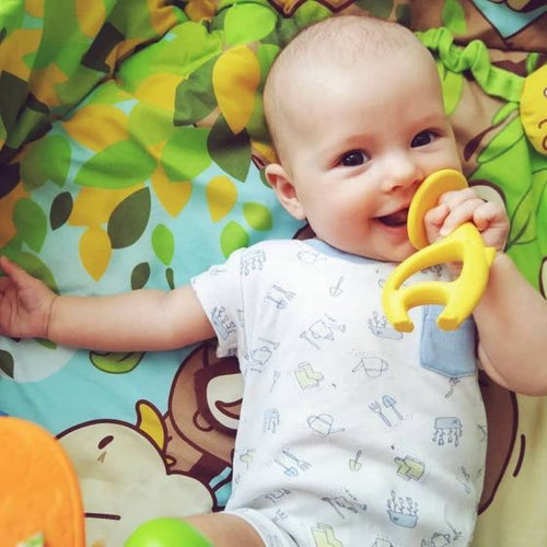 young baby lying on a colourful mat smiling and chewing on a teether