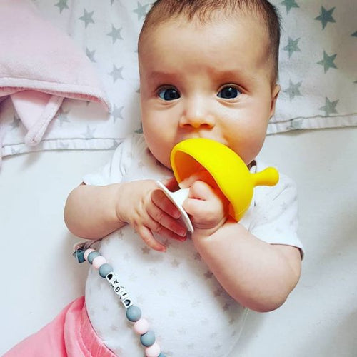 baby lying on blankets holding a yellow teether and chewing on it looking up