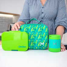 a person showing a go green yumbox, green pixel print insulated lunch bad and  and green insulated food jar on a white bench