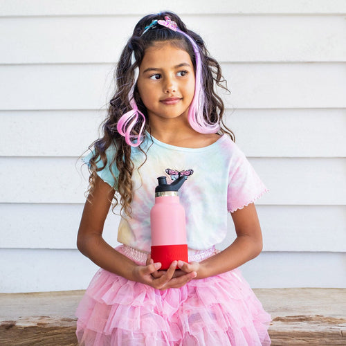 child in a pink tutu skirt, pastel the dye t-shirt, long dark hair with pink stripes added, holding a pale pink bottle with a red bumper