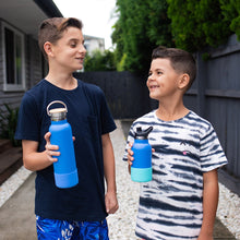 2 children standing outside in a driveway looking at each other holding blue drink bottles with blue and mint bumpers 