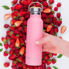 a hand holding a bold bright pink drink bottle in front of fresh strawberries - some are cut up, raspberries and green leaves