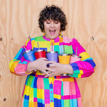 a person wearing a bright multi coloured dress holding five different colours of smoothie cups in front of a wooden background