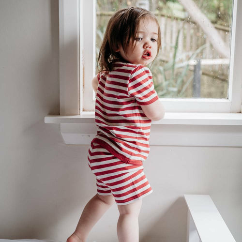 toddler standing on the bed in front of a window looking back into the room in red and white stripe t-shirt and short pyjamas