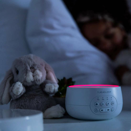 dark bedroom with person asleep, a soft bunny toy sitting beside the white noise machine with the red night light glowing
