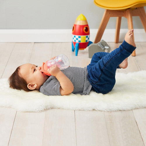 child lying down on sheepskin wear jean pants and a black and white stripe t-shirt drinking from a b.box watertermelon sippy cup, a wooden toy rocket and yellow chair with wooden legs is in the background